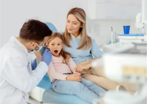 Pediatric dentist performing a dental exam for a young girl while her mother provides support.